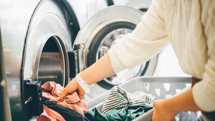 Interior Of Small Laundromat In Daylight. Close Up Female Holding Basket. Girl Loading Dirty Clothes Inside Drum And Closing Door.